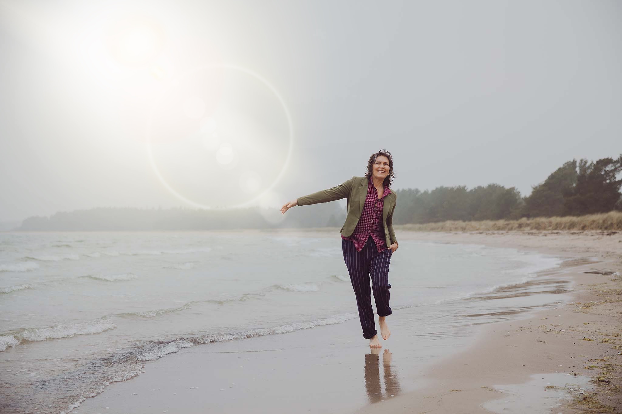 Vivianne vandrar på en sandstrand med havet stilla till vänster. En solstråle lyser ner på henne. Hon är barfota i blåbyxor och grönkavaj. Hon ser glad och svänger ut med vänster arm